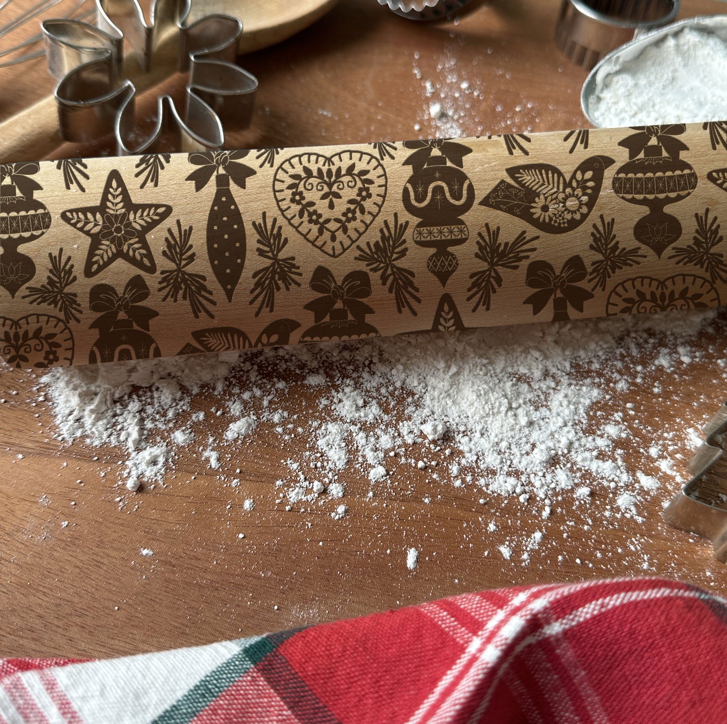 Decorative rolling pin with Christmas patterns on a wooden surface with flour and a plaid cloth.