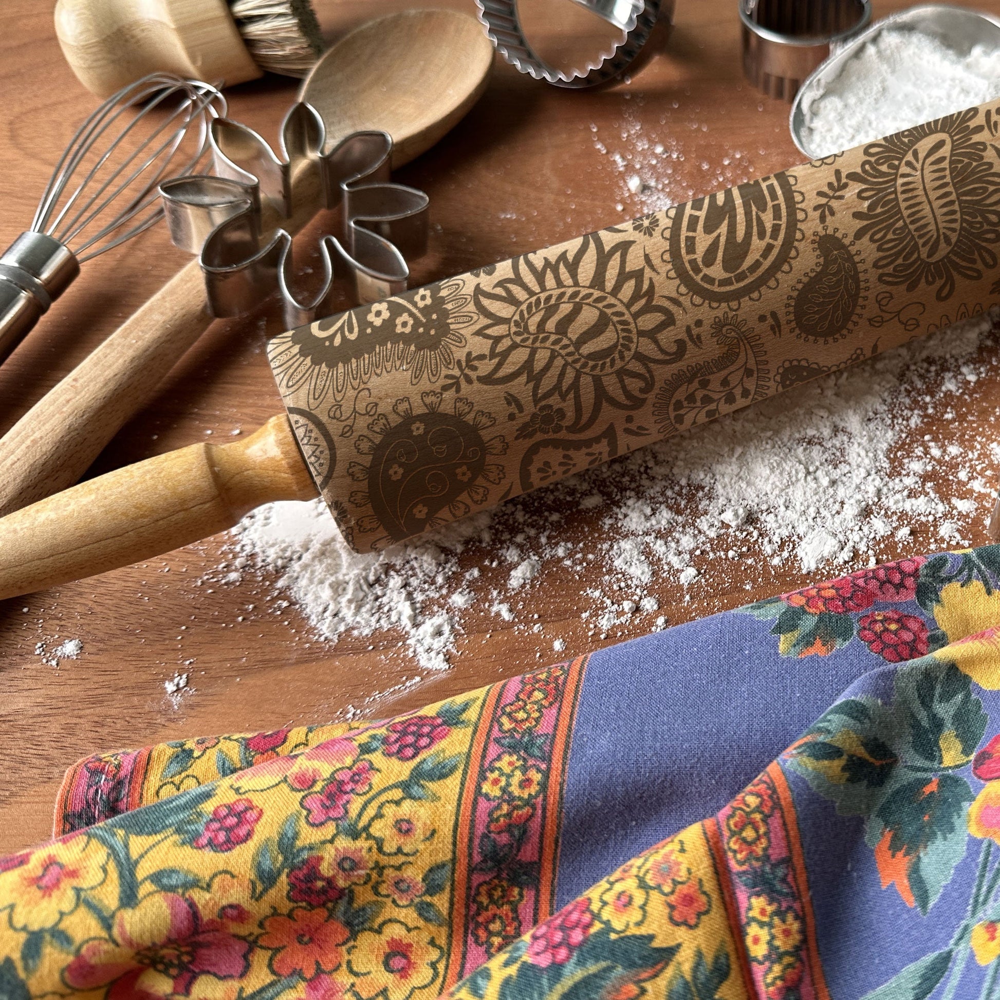 Decorative rolling pin with floral patterns on a wooden surface with kitchen utensils and a colorful towel.
