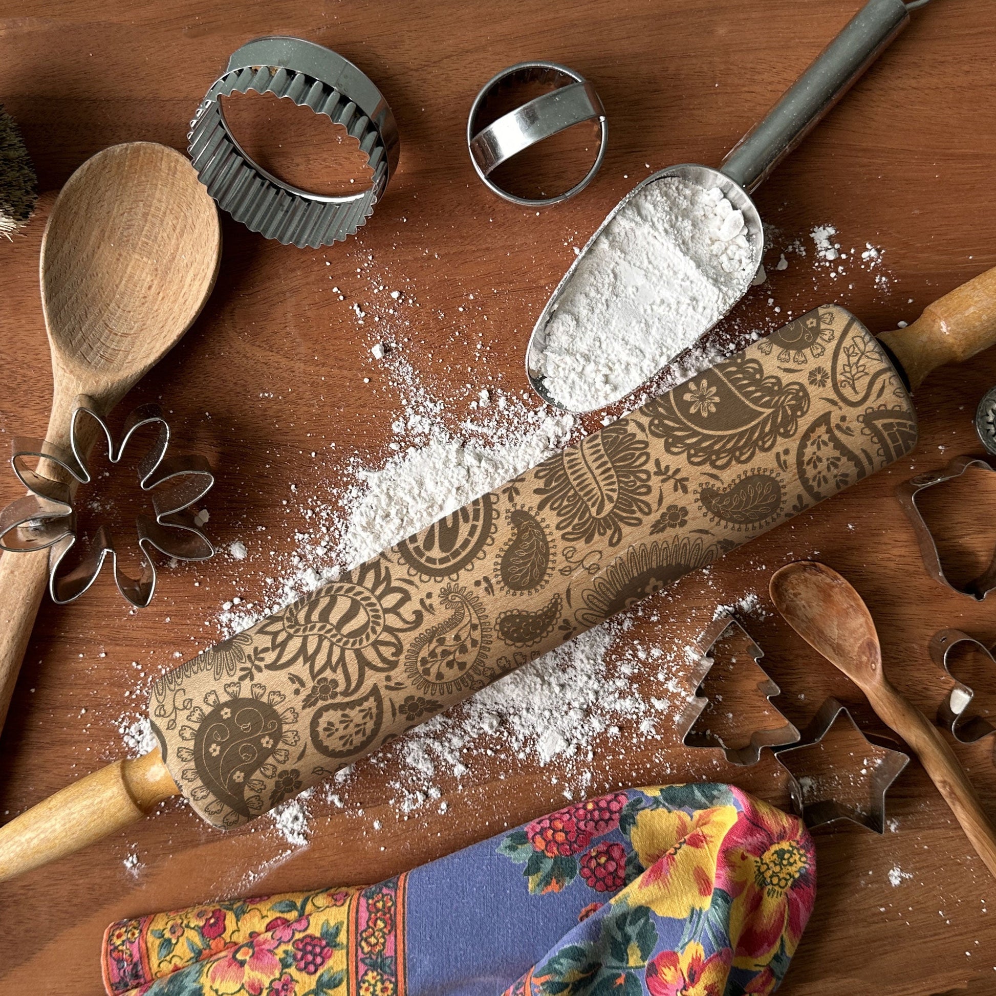 Wooden rolling pin with decorative patterns on a wooden surface with flour and cookie cutters.