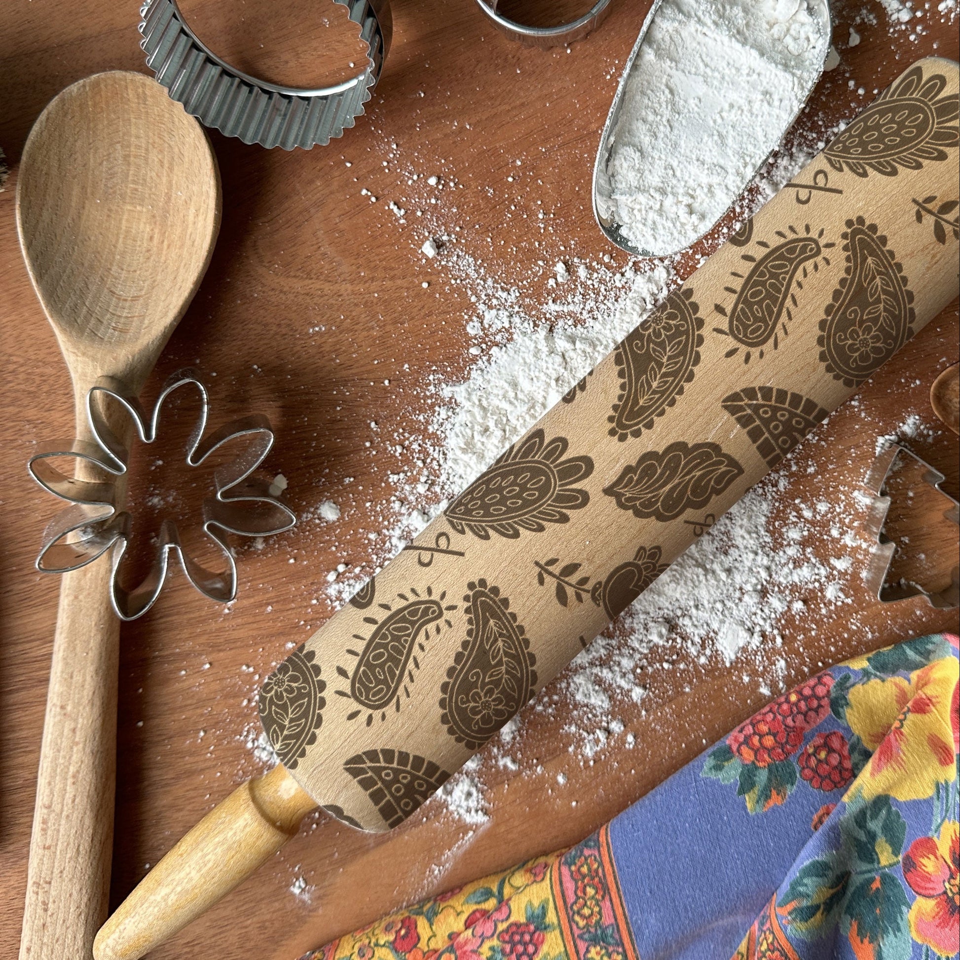 Baking tools including a wooden spoon, rolling pin with paisley patterns, and cookie cutters on a wooden surface.