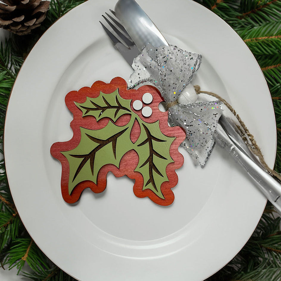 Holly Christmas ornament on a white plate with silverware, surrounded by greenery and pinecones.