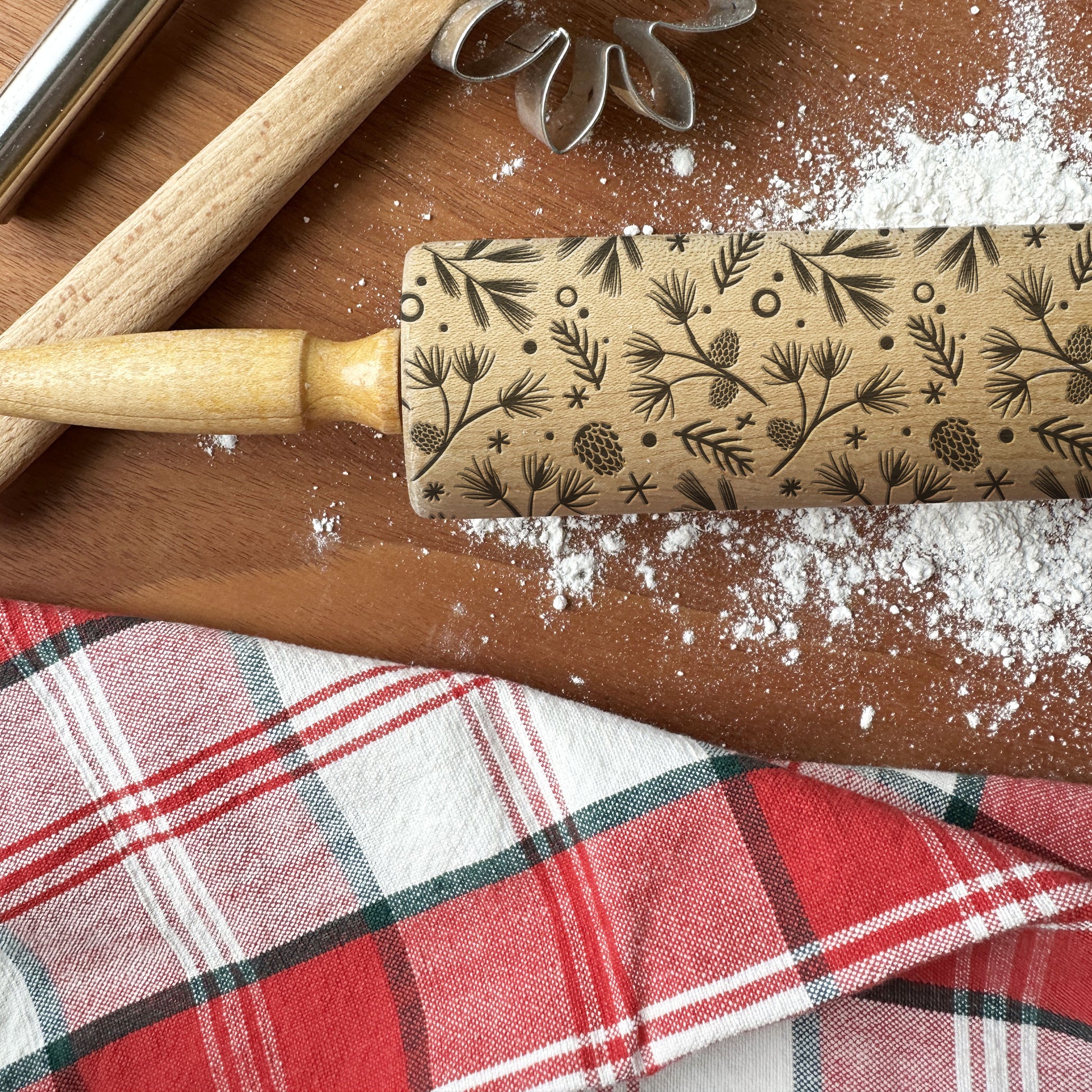 Decorative rolling pin with floral pattern on a wooden surface with flour and plaid fabric.