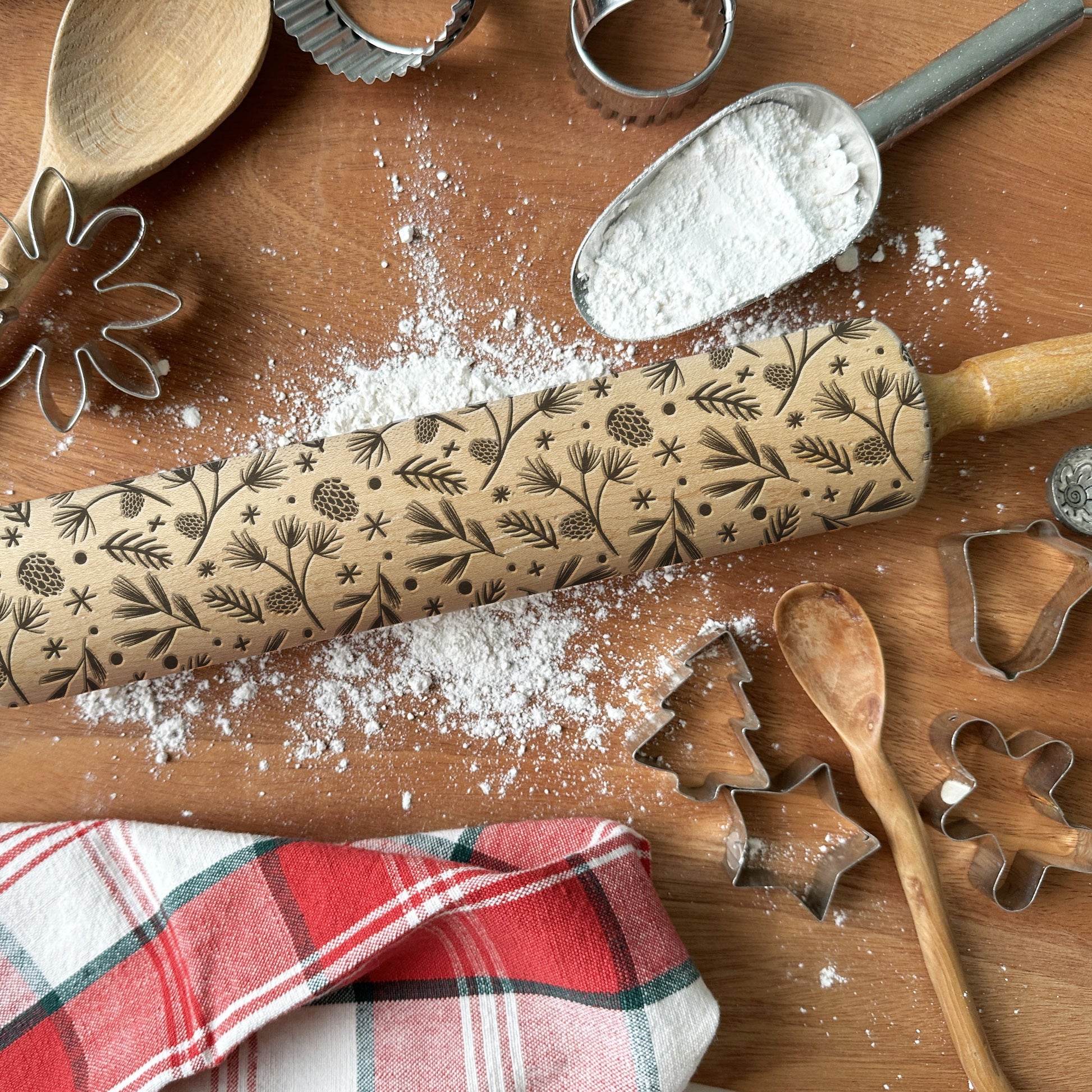 Wooden rolling pin with floral patterns on a wooden surface with flour, cookie cutters, and a plaid cloth.