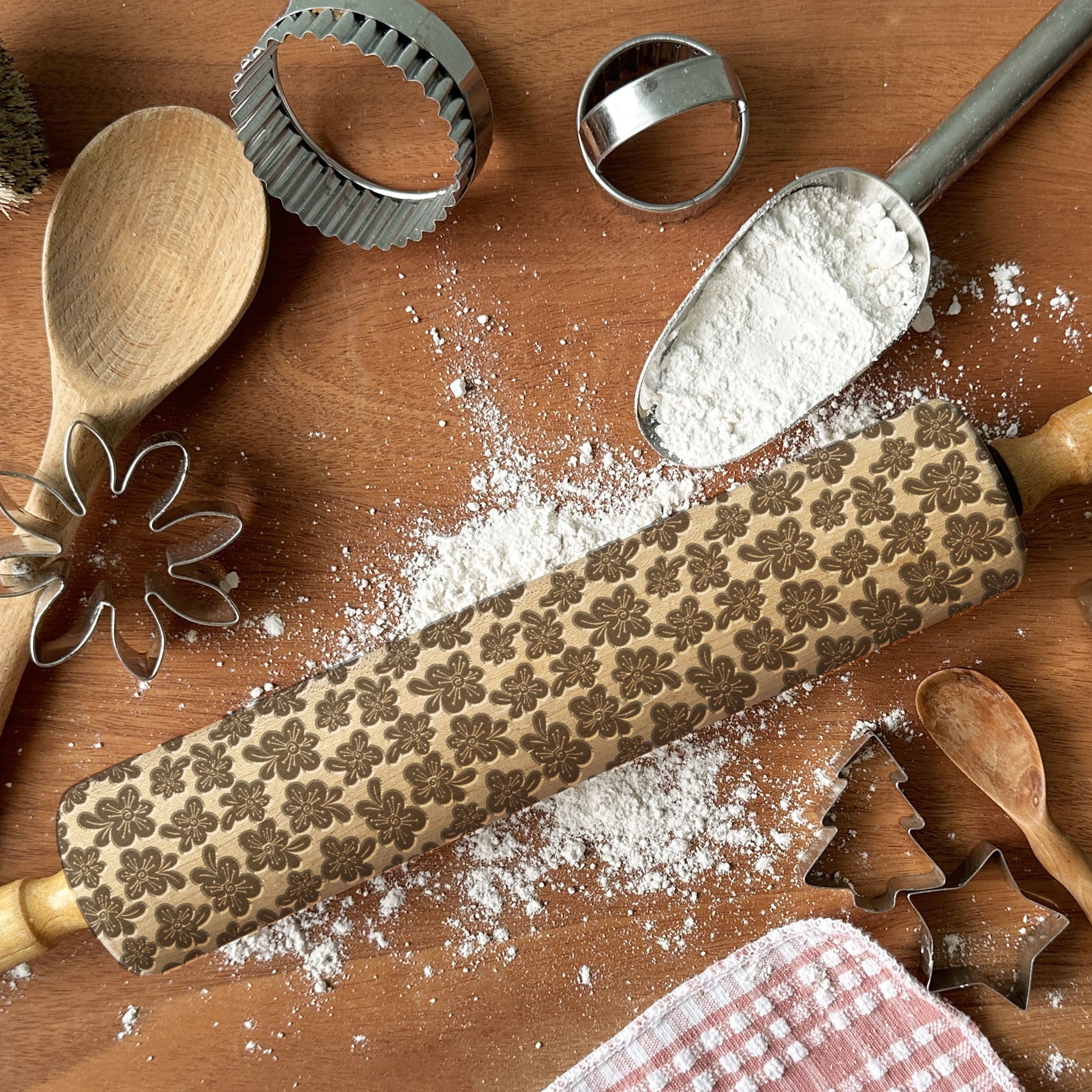Baking tools including a rolling pin with floral patterns, wooden spoons, and cookie cutters on a wooden surface with flour.