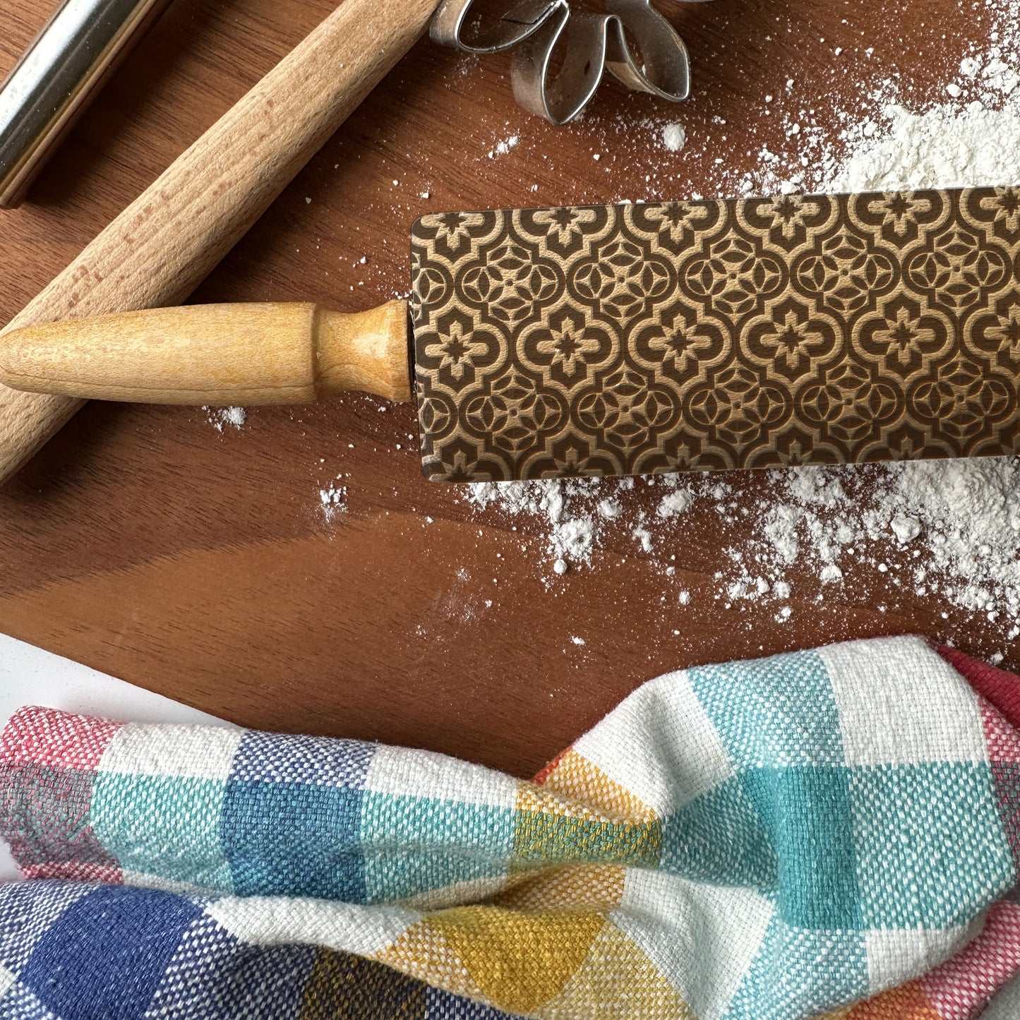 Decorative rolling pin with floral patterns on a wooden surface with flour and a checkered cloth.