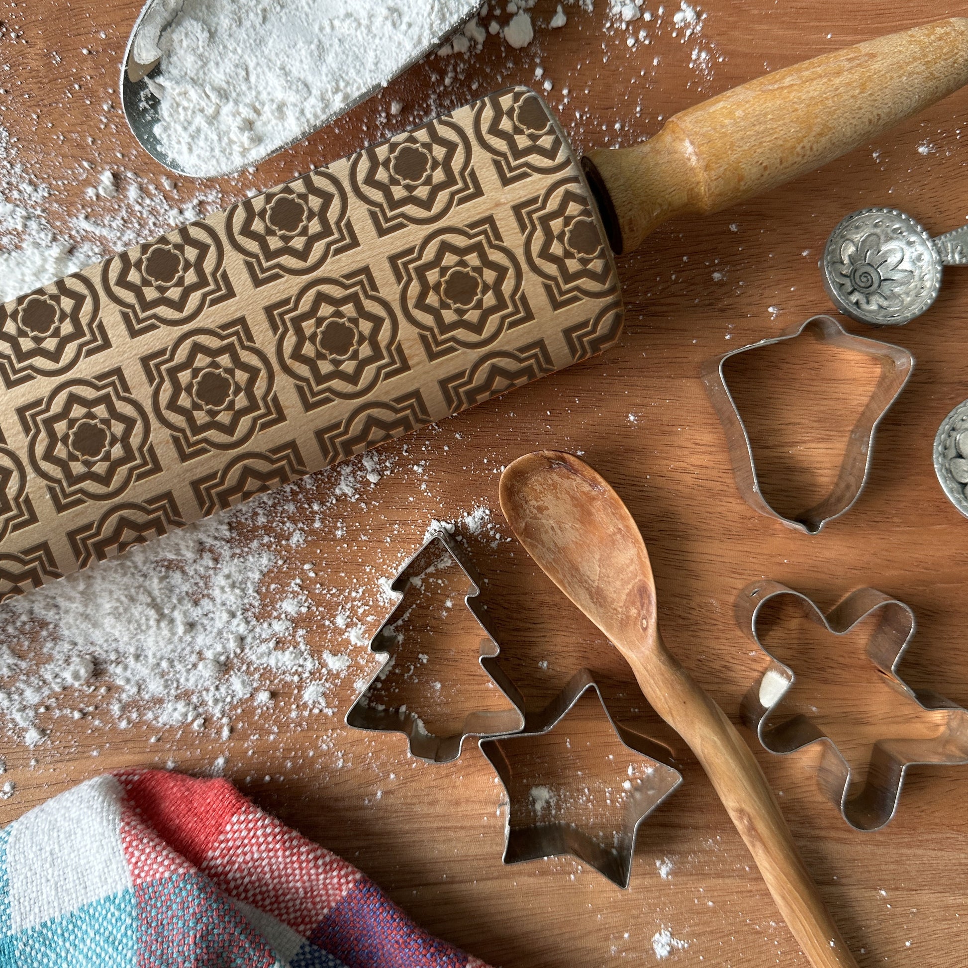 Baking tools including a rolling pin with embossed patterns, cookie cutters, and a wooden spoon on a wooden surface with flour.