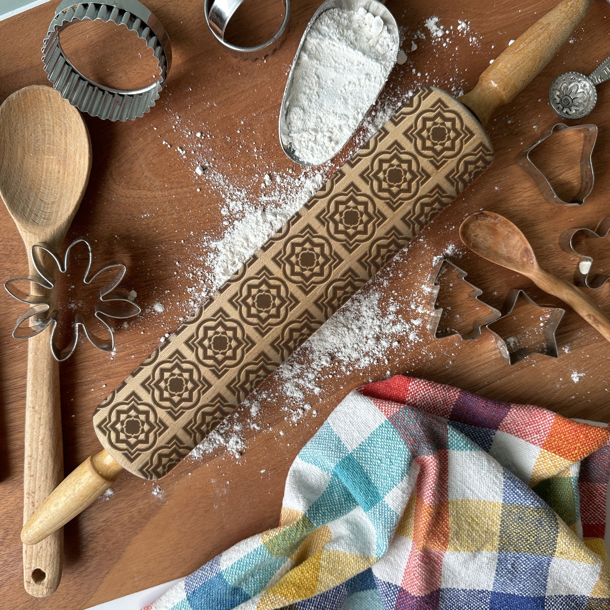 Decorative rolling pin with pattern, flour, wooden spoon, and cookie cutters on a wooden surface.