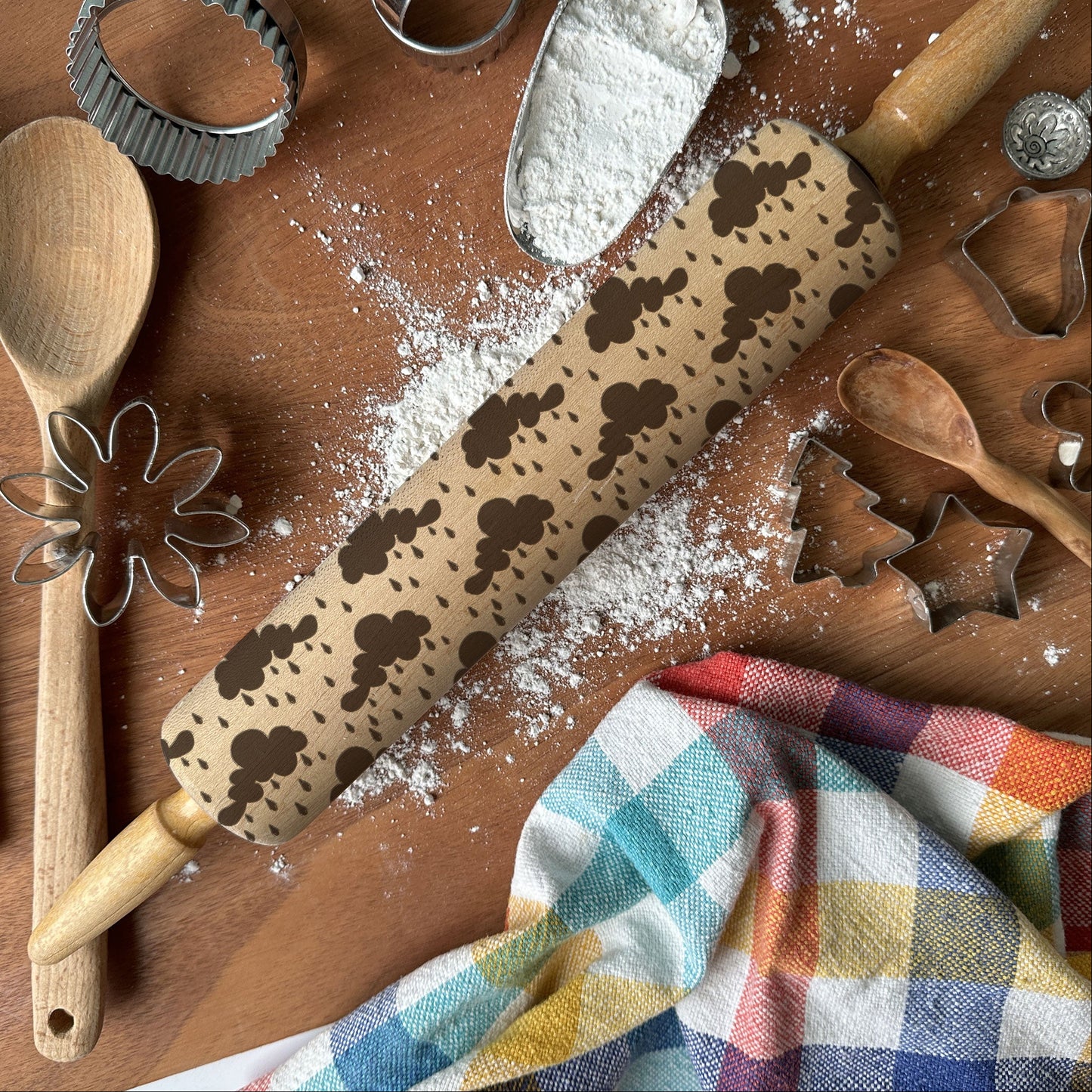 Baking tools including a rolling pin with cookie stamps, wooden spoon, whisk, and flour on a wooden surface.