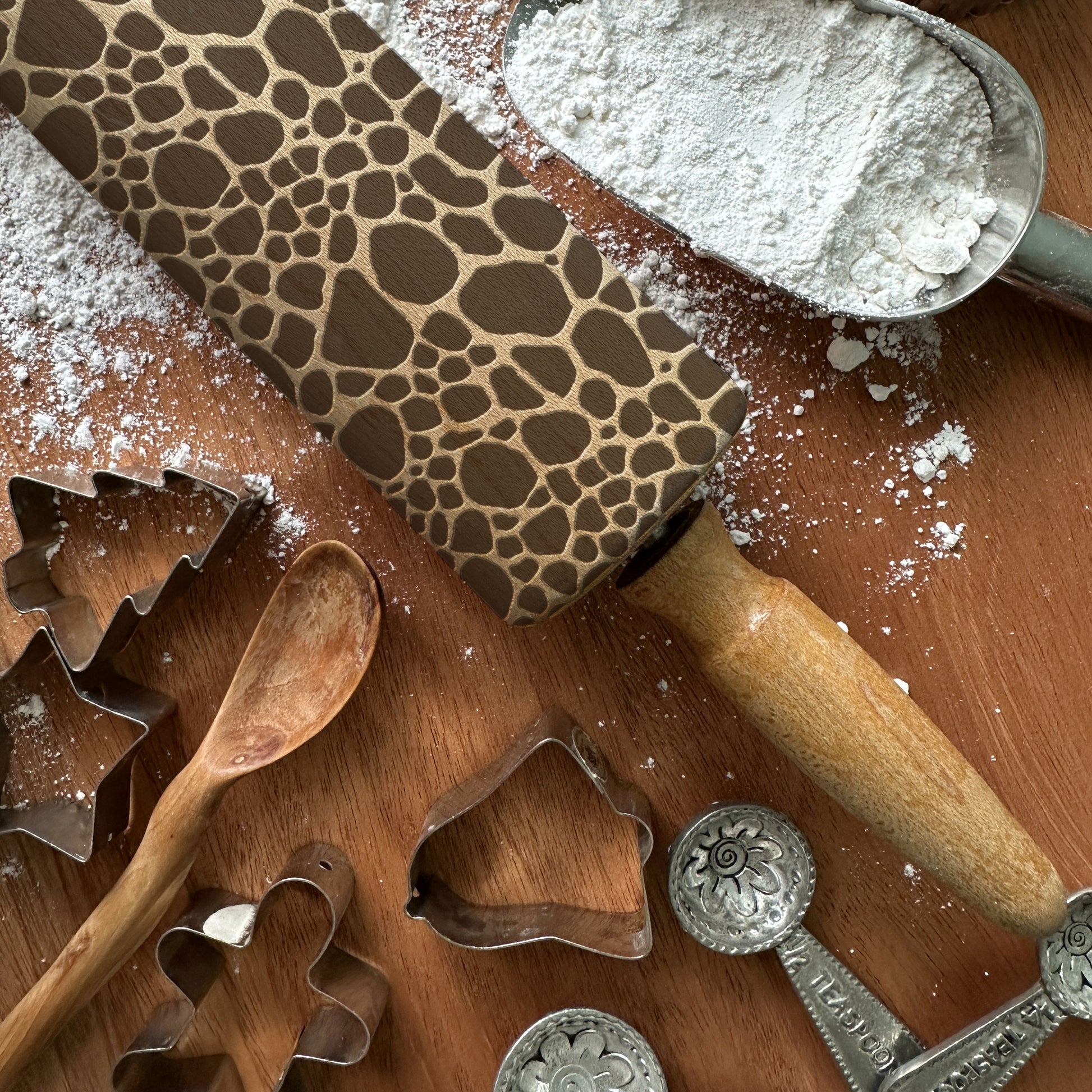 Wooden rolling pin with a pattern, flour, wooden spoon, and metal cookie cutters on a wooden surface.