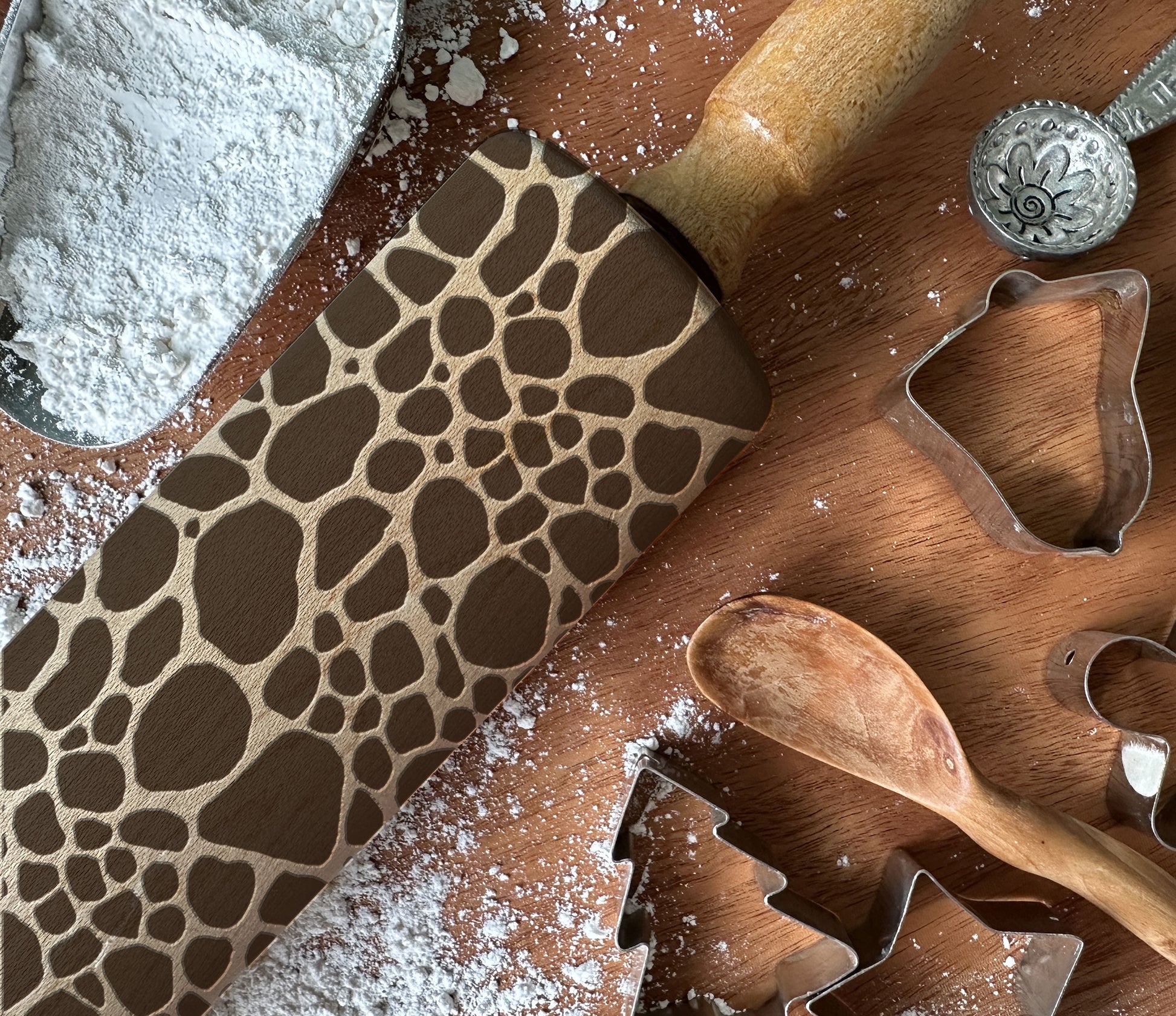 Baking tools including a patterned rolling pin, wooden spoon, and cookie cutters on a wooden surface with flour.