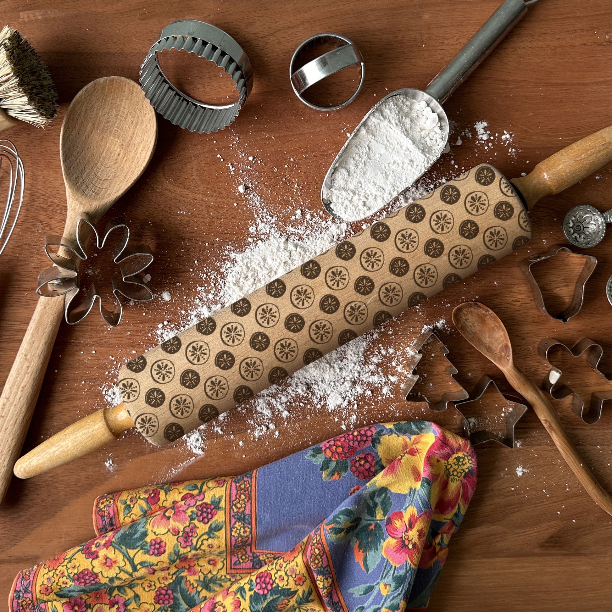 Baking tools including a rolling pin with patterned design, wooden spoons, and flour on a wooden surface.