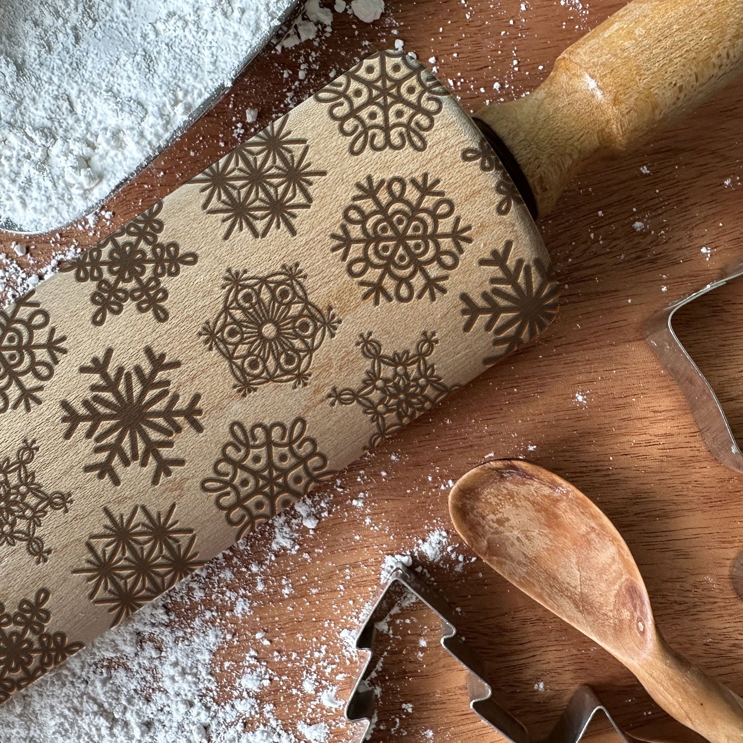 Wooden rolling pin with snowflake pattern on a wooden surface with flour and cookie cutters.