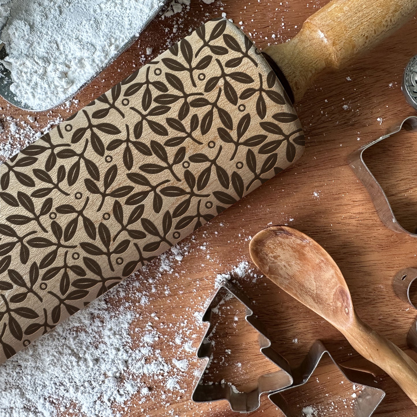 Wooden rolling pin with leaf pattern on a wooden surface with cookie cutters and flour.