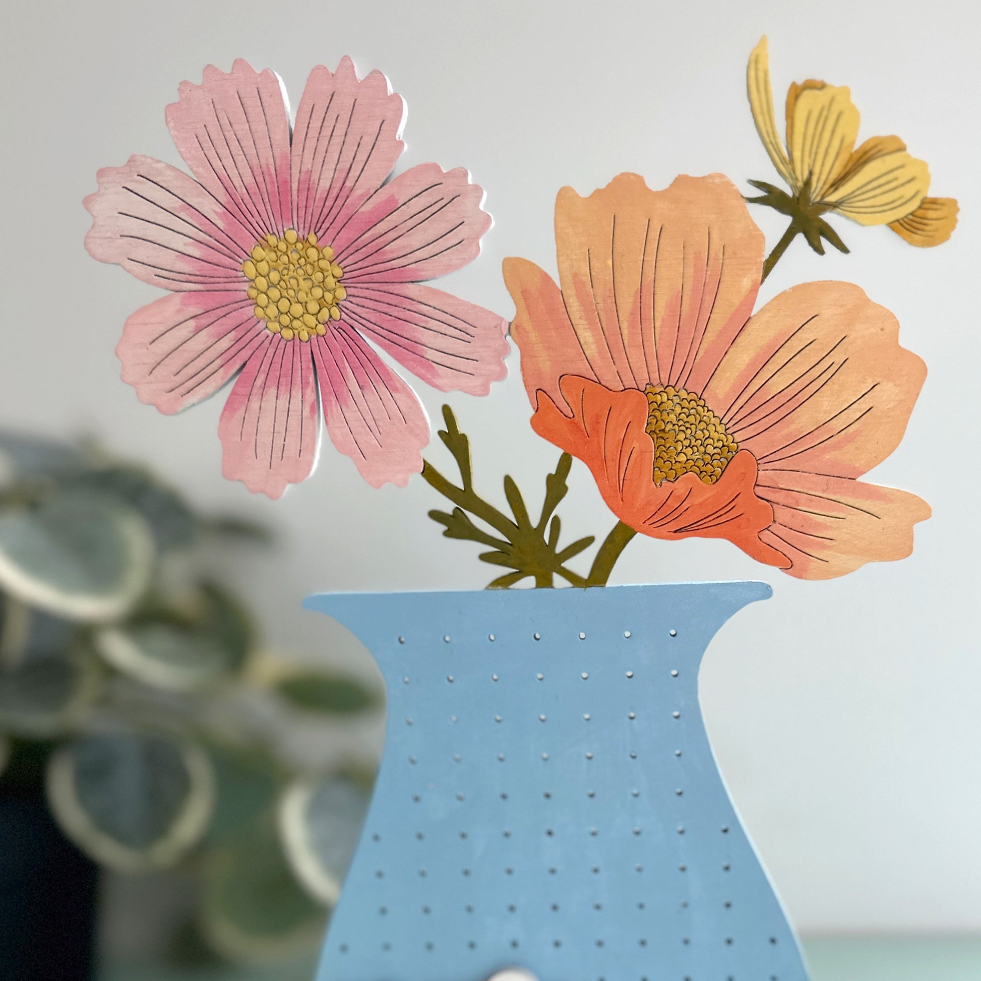 Decorative wall art with pink and orange flowers in a blue vase on a light gray background.