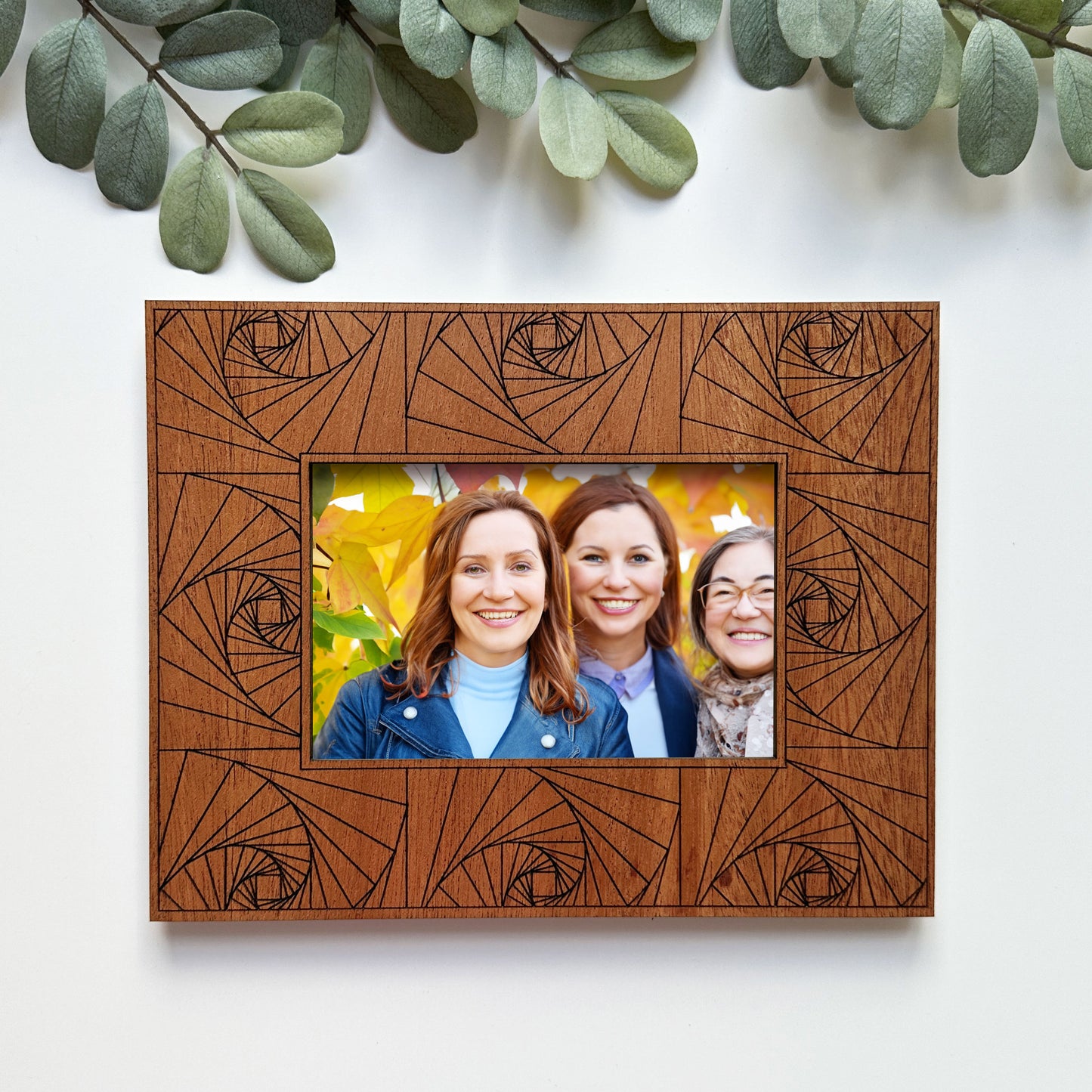 Wooden picture frame with geometric design holding a photo of three people, surrounded by green leaves.