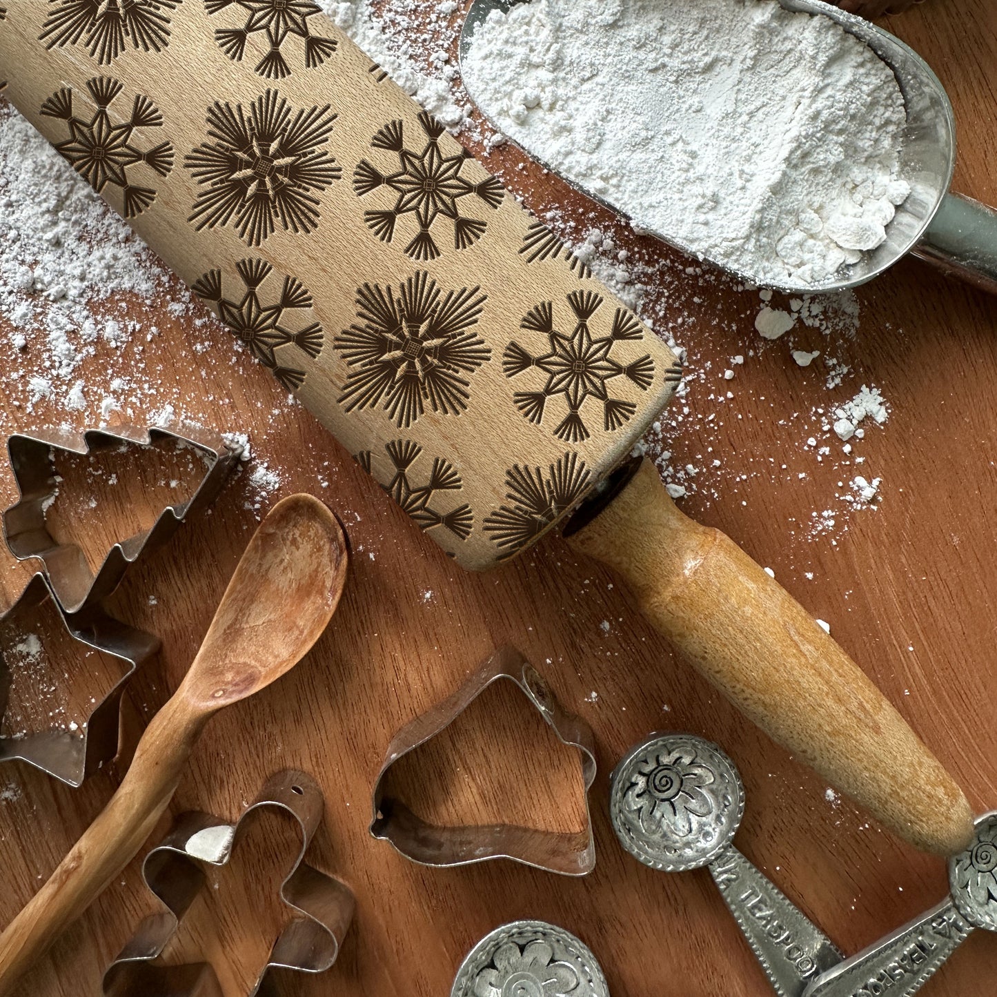 Rolling pin with snowflake patterns on a wooden surface with cookie cutters and flour.