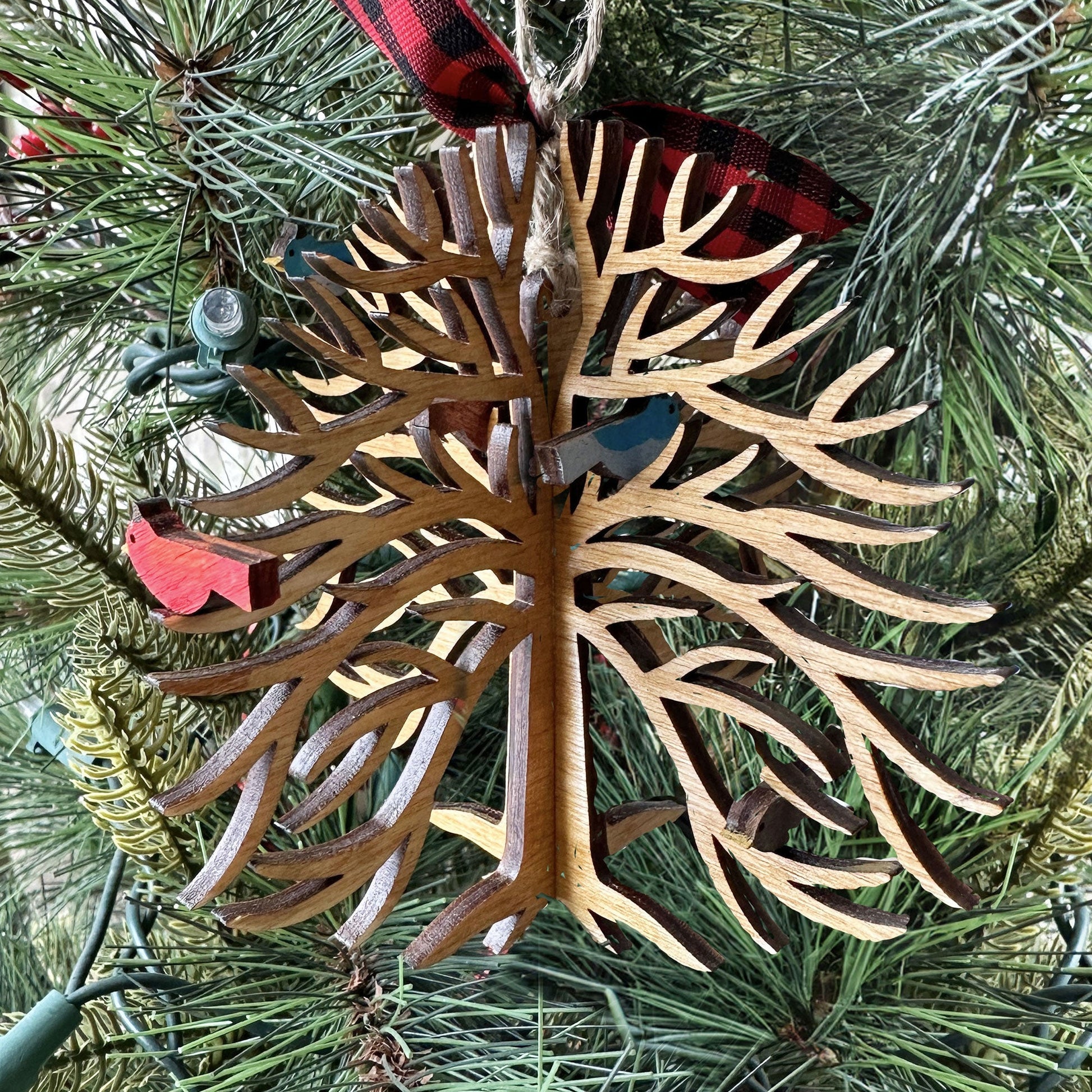 Decorative wooden ornament with a bird on a tree branch, hanging from a red and black plaid ribbon, against a Christmas tree background.