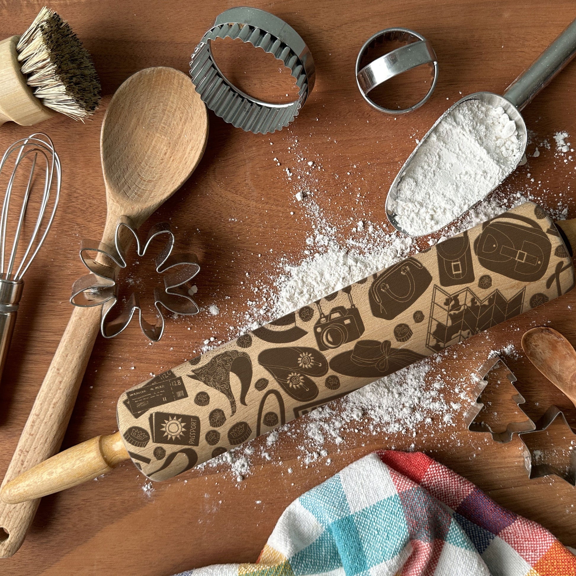 Baking tools including a rolling pin with decorative patterns, wooden spoon, whisk, and cookie cutters on a wooden surface.