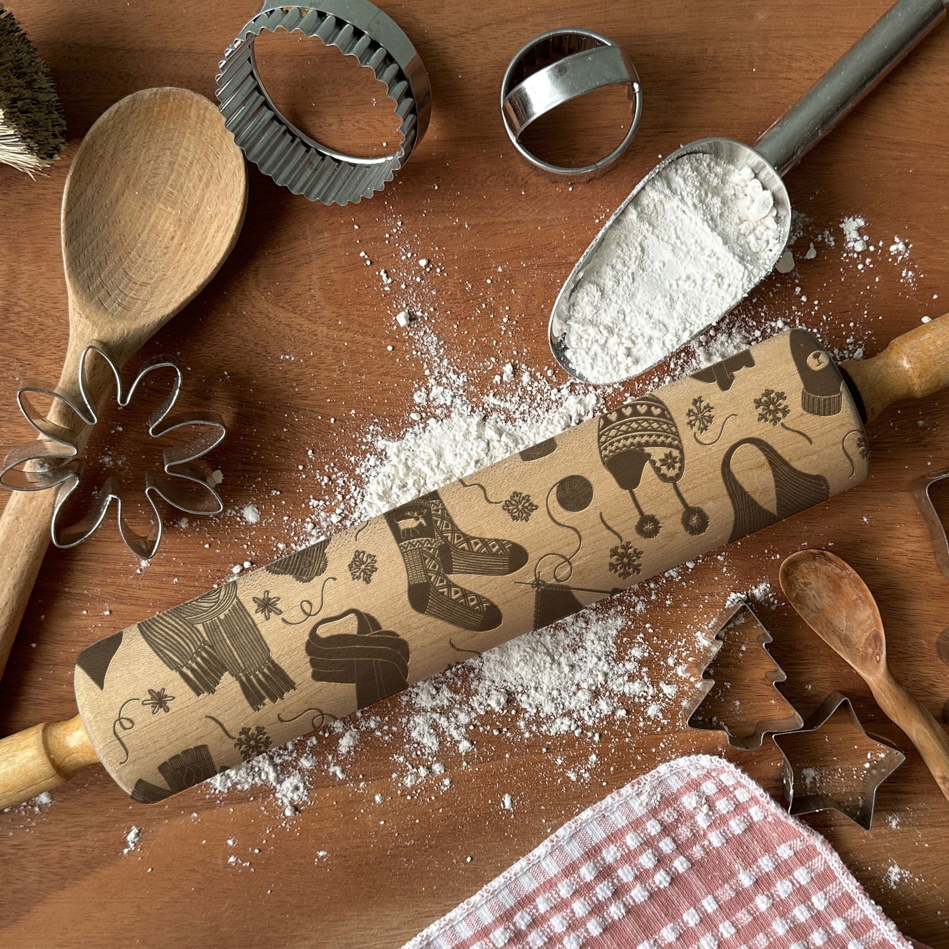 Baking tools including a wooden spoon, rolling pin with decorative patterns, and cookie cutters on a wooden surface with flour.