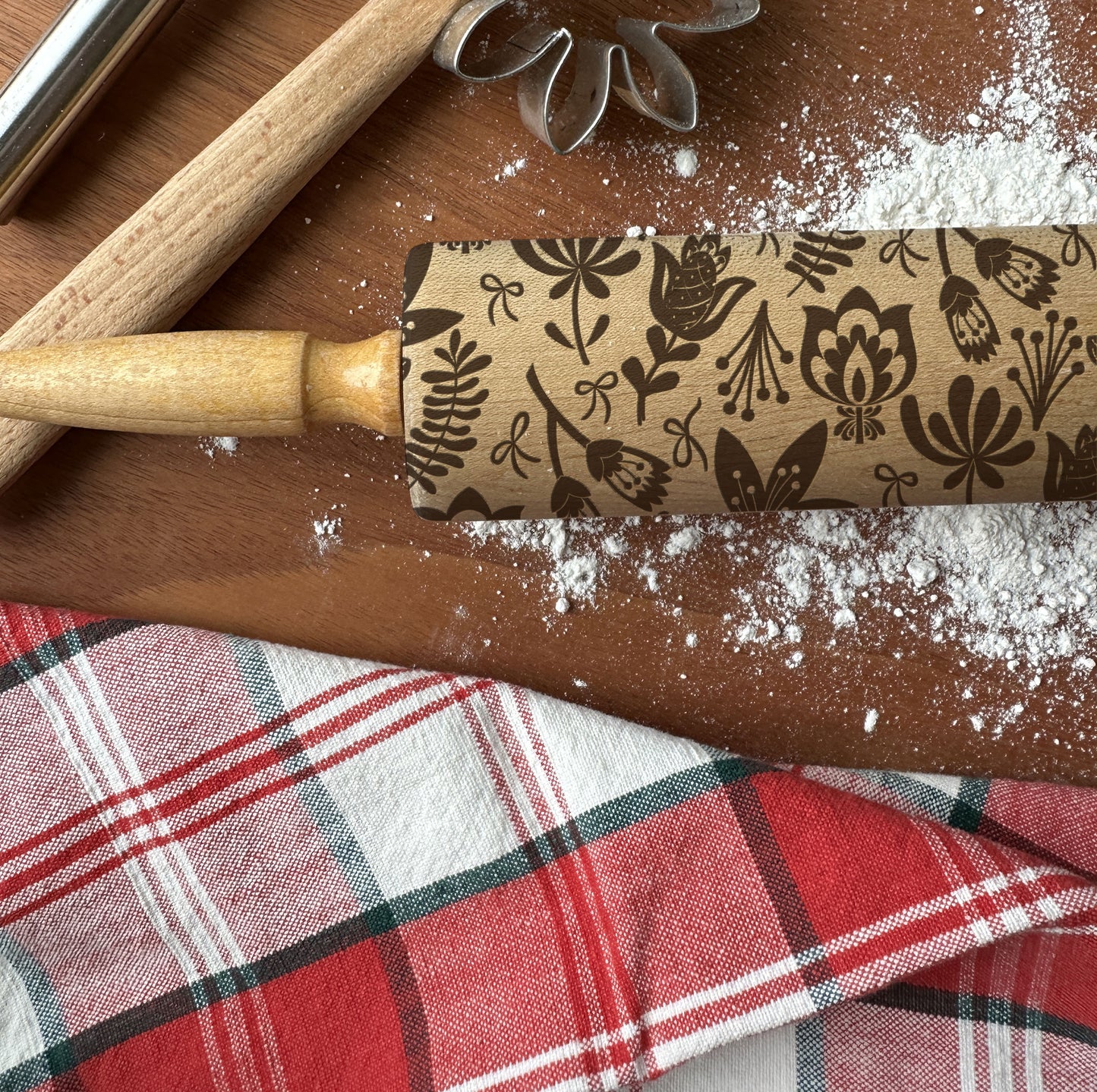 Decorative rolling pin with floral pattern on a wooden surface with flour and plaid fabric.