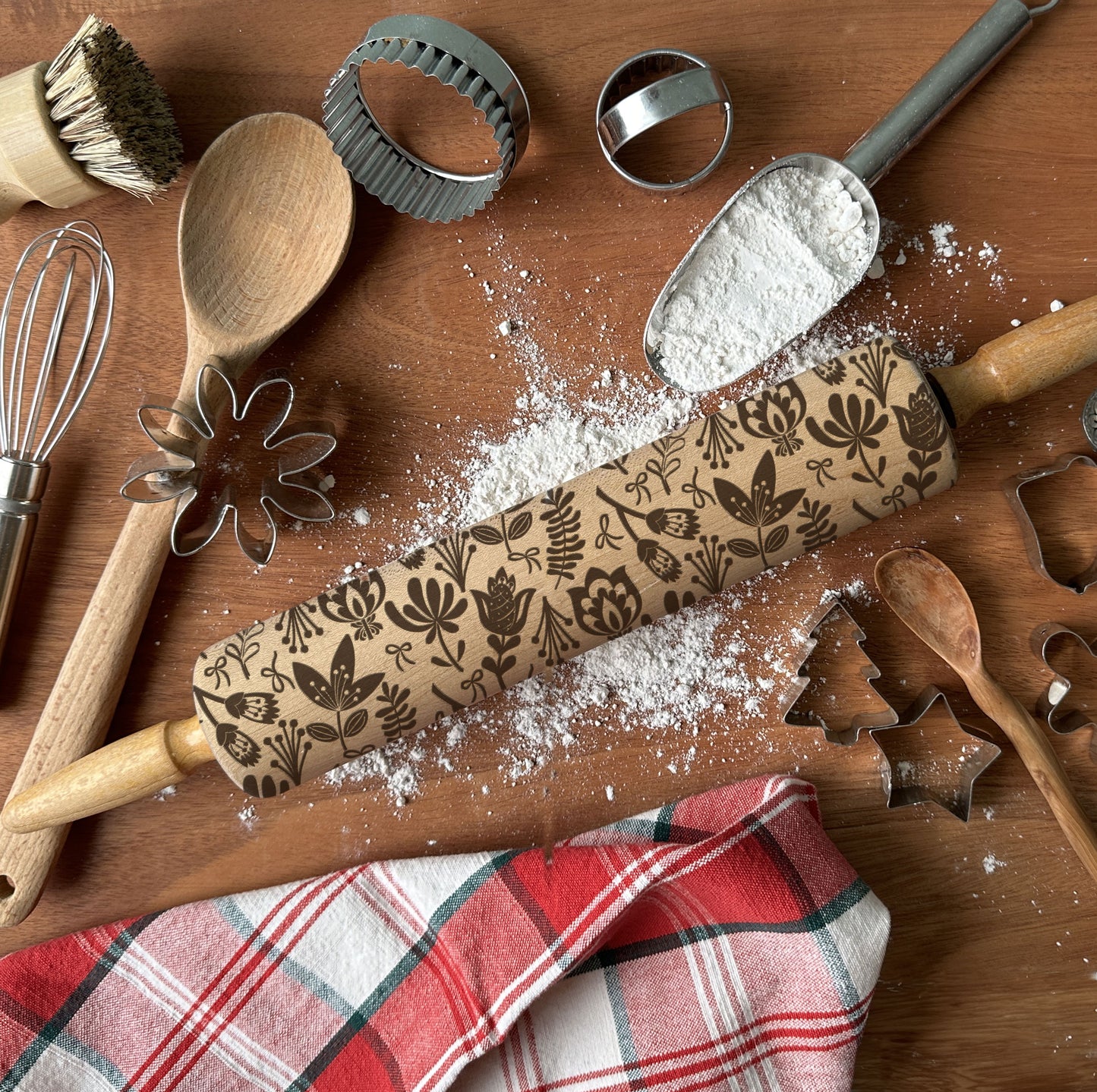 Baking tools including a rolling pin with embossed designs, wooden spoon, whisk, and cookie cutters on a wooden surface.