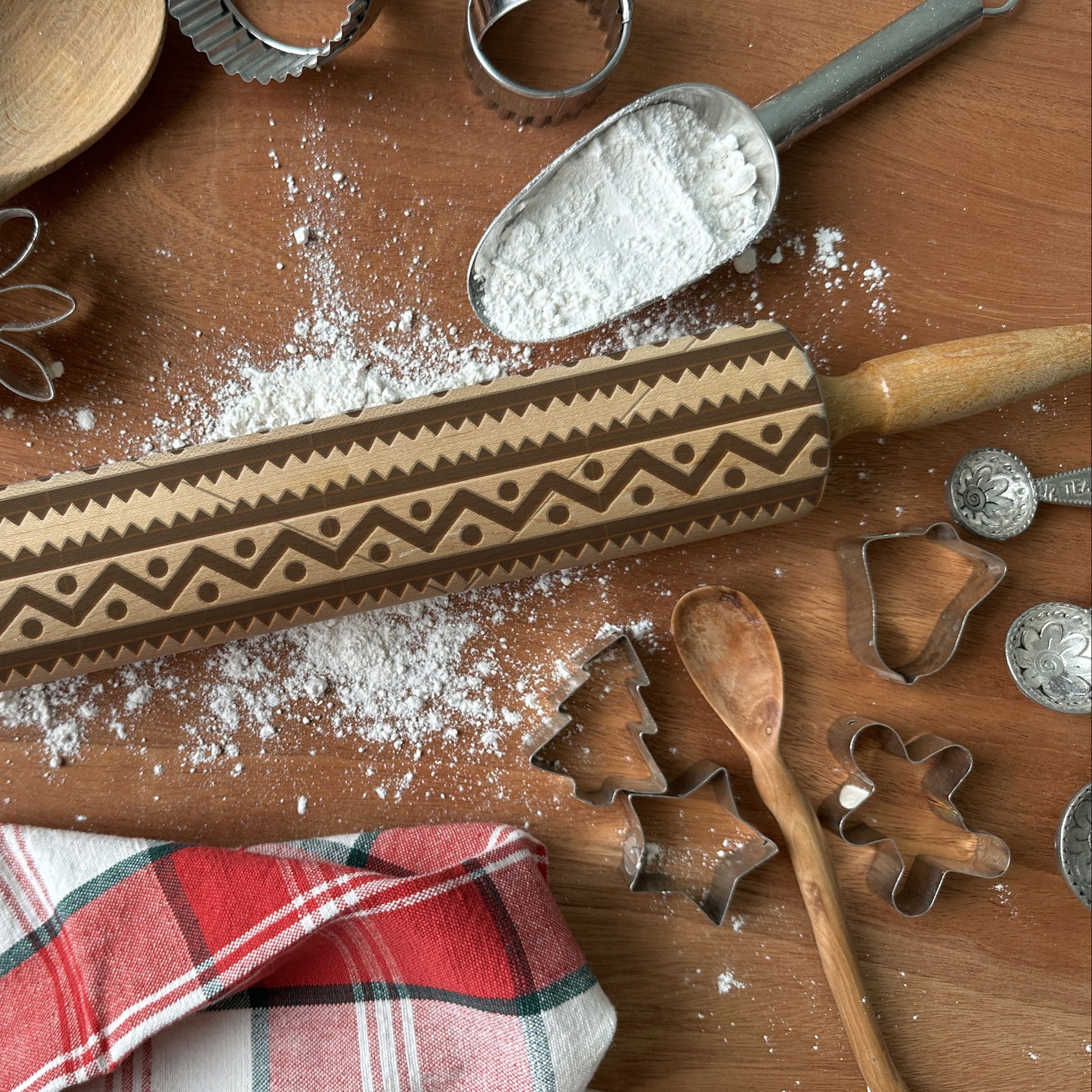 Wooden rolling pin with decorative patterns on a wooden surface with flour, cookie cutters, and a red plaid cloth.